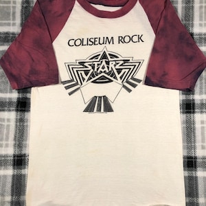 May include: A white and burgundy baseball-style t-shirt with the text "COLISEUM ROCK" above a black and white star graphic. The shirt has burgundy sleeves and a white body.