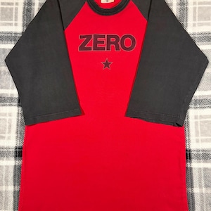 May include: A red and gray raglan-style t-shirt. The shirt features a red body with gray sleeves. The word "ZERO" is printed across the chest in a dark red font, with a star below it. The shirt has a crew neck.
