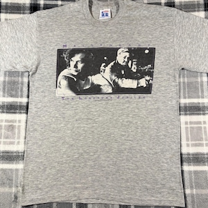 May include: A heather gray t-shirt featuring a black and white image of two men. The text "MELLENCAMP" is above the image, and "THE LONESOME JUBILEE" is below it in purple lettering. The shirt has a crew neck and short sleeves.