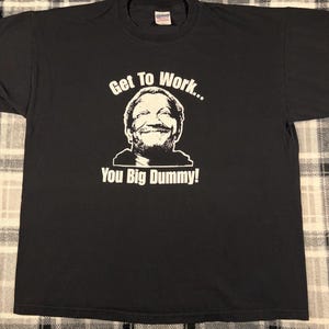 May include: A black short-sleeved t-shirt featuring a white graphic print of a man's face. Above the face is the text "Get To Work..." and below the face is the text "You Big Dummy!" The shirt has a crew neckline and a standard fit.