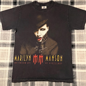 May include: Black t-shirt featuring a portrait of Marilyn Manson with red lipstick and a black hat. The text "MARILYN MANSON" is printed in yellow and red, with "THE GOLDEN AGE OF GROTESQUE" below.