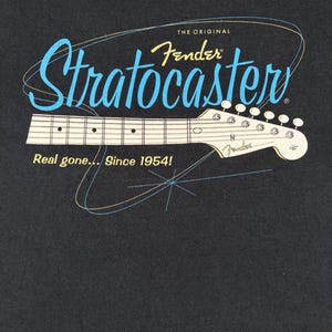 May include: A black t-shirt featuring a graphic of a Fender Stratocaster guitar neck. The text reads THE ORIGINAL Fender Stratocaster and Real gone... Since 1954! The design includes light blue and yellow line art accents on a dark background.