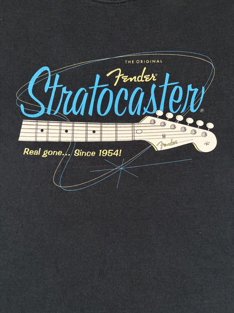 May include: A black t-shirt featuring a graphic of a Fender Stratocaster guitar neck. The text reads THE ORIGINAL Fender Stratocaster and Real gone... Since 1954! The design includes light blue and yellow line art accents on a dark background.