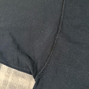 May include: A close-up view of a black fabric garment, specifically focusing on the underarm area where the sleeve meets the body. The image displays the dark textile texture, the vertical seam construction, and the stitching detail of the clothing item.