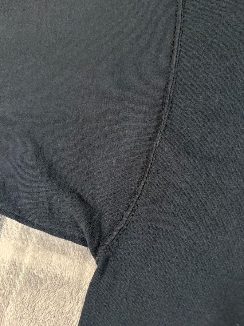 May include: A close-up view of a black fabric garment, specifically focusing on the underarm area where the sleeve meets the body. The image displays the dark textile texture, the vertical seam construction, and the stitching detail of the clothing item.