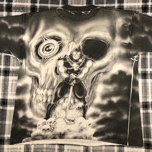 May include: Black and white graphic t-shirt featuring a large skull with a menacing figure holding a weapon. The figure is standing on a pile of skulls. The shirt has a crew neck and short sleeves.