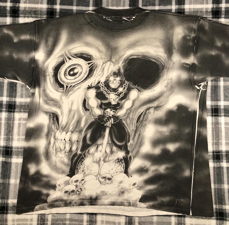 May include: Black and white graphic t-shirt featuring a large skull with a menacing figure holding a weapon. The figure is standing on a pile of skulls. The shirt has a crew neck and short sleeves.