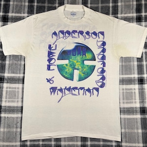 May include: Cream-colored t-shirt featuring a graphic design with the words "Anderson, Bruford, Wakeman, Howe" in purple. The central design is a blue and green circular image with a cross-like shape.
