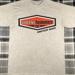 May include: A heather gray t-shirt featuring the band name "VIOLENT femmes" in a red and black hexagon design. Below the logo, the text reads "Since 1981" and "american music". The shirt has a crew neck and short sleeves.