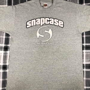 May include: Gray t-shirt with a white graphic of the word "snapcase" in an arc above a stylized letter "S" with the year "MCMXCVII" below it.