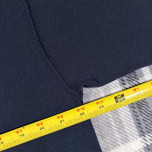 May include: A dark navy blue fabric garment featuring a visible seam along the armhole. A yellow measuring tape is positioned across the fabric, showing a measurement extending from 18 inches to 25 inches along the edge of the textile material.