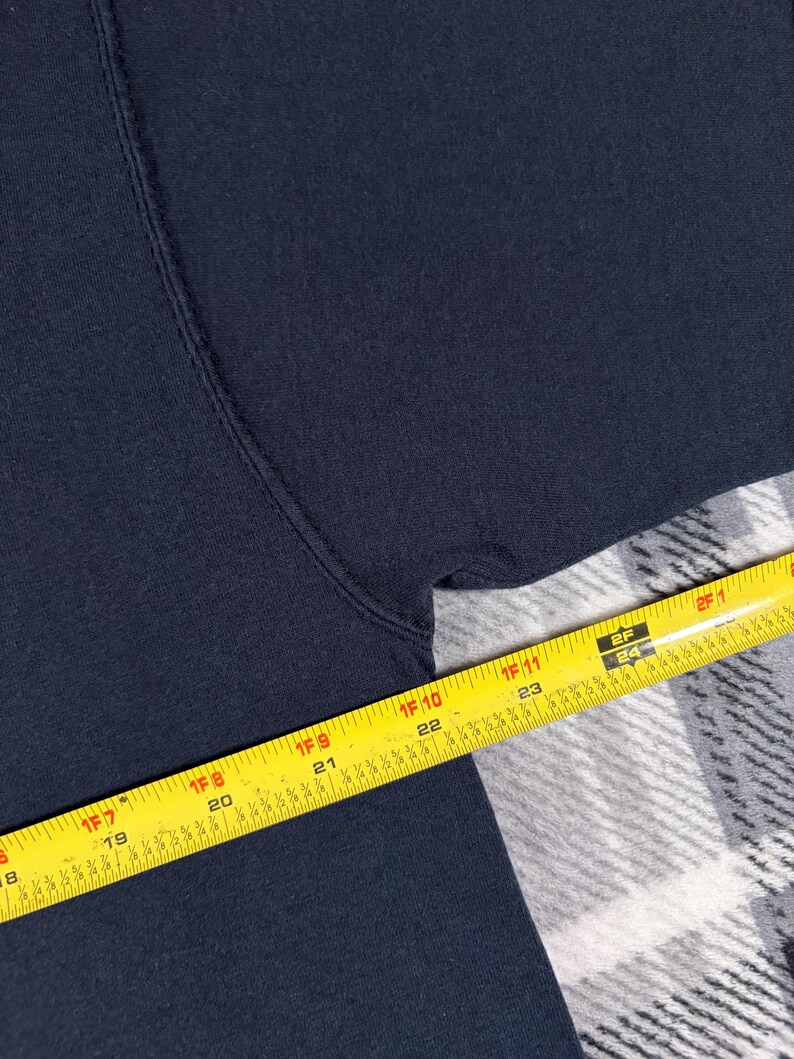 May include: A dark navy blue fabric garment featuring a visible seam along the armhole. A yellow measuring tape is positioned across the fabric, showing a measurement extending from 18 inches to 25 inches along the edge of the textile material.