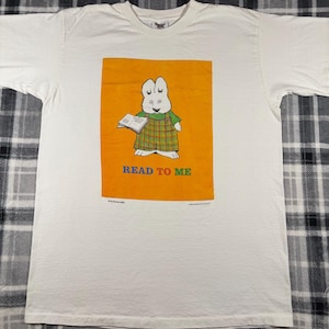May include: White t-shirt featuring a cartoon rabbit character holding a book on an orange background. The rabbit wears a green plaid outfit. The text "READ TO ME" is below the image in colorful letters. A vintage children's apparel item.