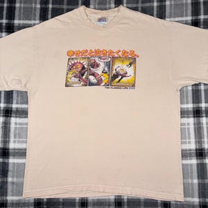 May include: Beige t-shirt featuring a comic strip design with Japanese text and the words "WHAT!!" in a speech bubble. The graphic includes images of figures in action. The text "THE FLAMING LIPS 2002" is printed below the comic strip.