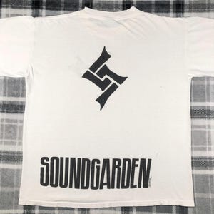 May include: White t-shirt featuring a black geometric design and the band name "SOUNDGARDEN" printed in bold, black letters. The shirt has short sleeves and a crew neck.