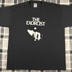 May include: Dark gray t-shirt featuring the text "THE EXORCIST" in white block letters above a white graphic. The graphic appears to be a silhouette of a building and people.