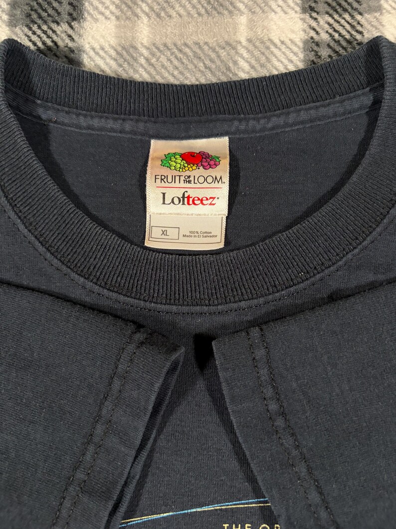 May include: A solid dark navy blue t-shirt with a ribbed crew neckline. The interior neck label features the Fruit of the Loom logo, the text Lofteez, and a size XL tag indicating 100% cotton construction, with the origin marked as El Salvador.