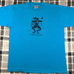 May include: A turquoise blue t-shirt with a crew neck. The shirt features a dark blue graphic of a figure with a crescent moon, the number 10, and the text "1993 Finding the God Within."