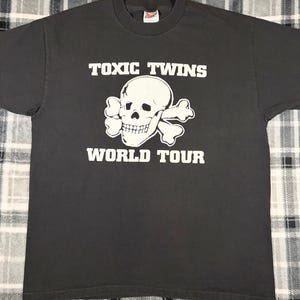 May include: Dark gray t-shirt with the text "TOXIC TWINS WORLD TOUR" in white block letters. A white skull and crossbones graphic is centered below the text. The shirt is made of a soft material.
