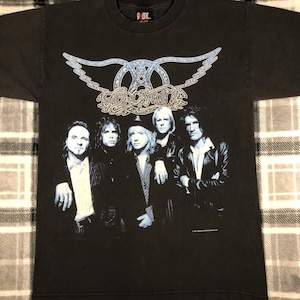 May include: Black t-shirt with a blue and white Aerosmith logo and a photo of the band members. The band members are wearing black clothing and are standing in front of a black background. The text "Aerosmith" is written in a stylized font.