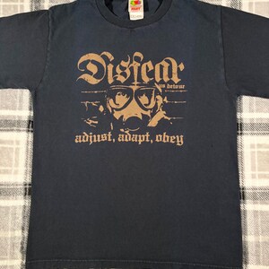 May include: A dark navy blue short-sleeve t-shirt featuring a tan graphic print. The design displays the text "Disfear" above an illustration of a person wearing a gas mask and goggles, with the text "us detour" and "adjust, adapt, obey" below.