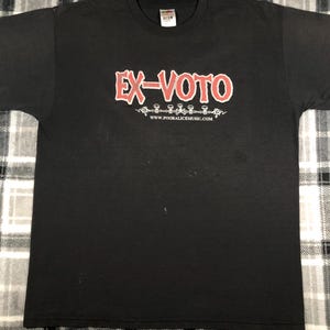 May include: Black t-shirt with the text "EX-VOTO" in red, outlined in white. Below the text is a decorative design and the website address www.pooralicemusic.com. The shirt has a crew neck and short sleeves.
