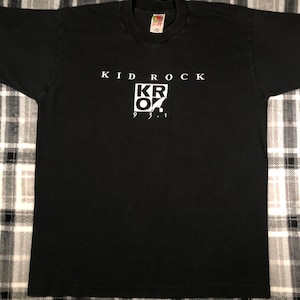 May include: Black t-shirt with the text "KID ROCK" above a white square containing "KR OK" and the numbers "9 3 1". The shirt is made of a soft material and has a crew neck.