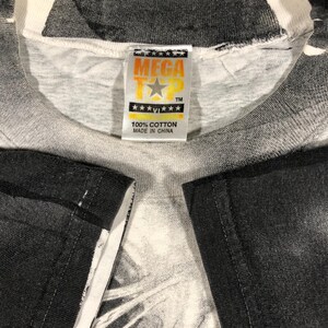May include: Close-up of a white and gray t-shirt with a graphic design. The shirt has a tag that reads "MEGA TEE" with a star logo and "100% COTTON MADE IN CHINA". The shirt's sleeves are black.