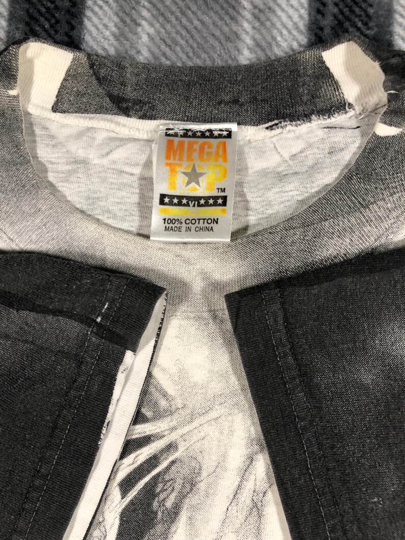 May include: Close-up of a white and gray t-shirt with a graphic design. The shirt has a tag that reads "MEGA TEE" with a star logo and "100% COTTON MADE IN CHINA". The shirt's sleeves are black.