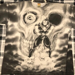 May include: A black and white graphic t-shirt featuring a large skull with a menacing figure holding a weapon. The figure is standing on a pile of skulls. The shirt has a crew neck and short sleeves.