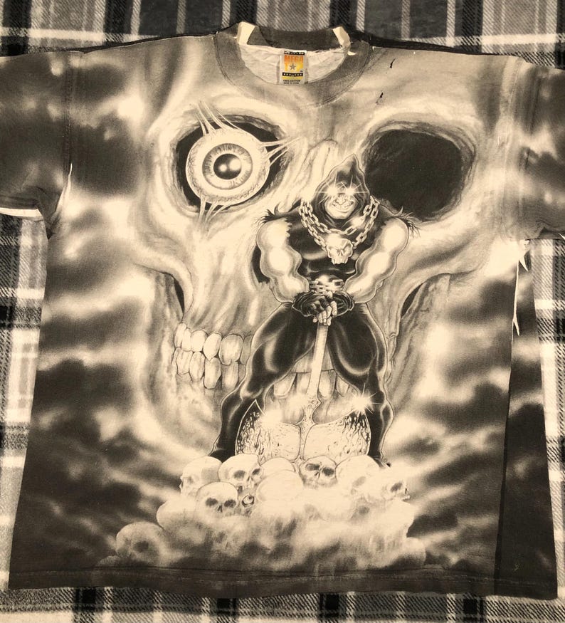 May include: A black and white graphic t-shirt featuring a large skull with a menacing figure holding a weapon. The figure is standing on a pile of skulls. The shirt has a crew neck and short sleeves.
