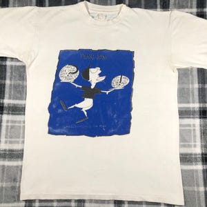 May include: Off-white t-shirt featuring a blue square graphic with the band name "Pearl Jam" and a cartoon figure. The figure is black and white, holding two circular objects with text. The shirt is a vintage band tee.