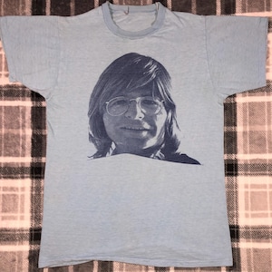 May include: A light blue t-shirt with a vintage print of a man with glasses and long hair. The man is smiling and looking directly at the viewer.