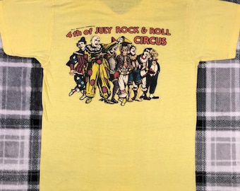 Puede incluir: Camiseta amarilla con un gráfico de un grupo de payasos frente a una carpa a rayas rojas y blancas. El texto "4th of JULY ROCK & ROLL CIRCUS" está impreso sobre los payasos.