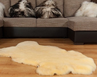 May include: A cream-colored sheepskin rug with a natural, fluffy texture. The rug is laid out on a wooden floor in front of a brown sectional sofa with white and black sheepskin throw pillows.