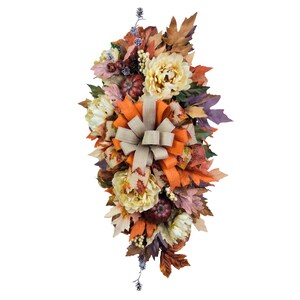 May include: A fall-themed floral arrangement featuring faux pumpkins, cream-colored peonies, and colorful leaves in shades of orange, brown, and purple. A large tan bow is centered in the design, creating a cascading effect.