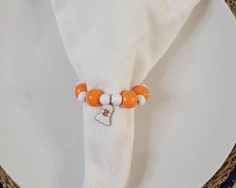 May include: A napkin ring featuring alternating orange and white spherical beads strung together. A small, white, ghost-shaped charm with a red accent is attached to the center of the beaded ring, which is wrapped around a folded white cloth napkin.