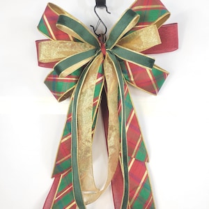 May include: A decorative bow featuring a red and green plaid pattern with gold metallic accents. The bow is constructed from multiple layers of ribbon, including solid dark green velvet-textured ribbon and shimmering gold fabric streamers.
