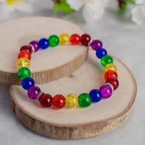 Könnte beinhalten: Ein regenbogenfarbenes Perlenarmband mit roten, orangefarbenen, gelben, grünen, blauen und violetten Perlen. Das Armband wird auf einer Holzscheibe präsentiert, mit weißen Blumen im Hintergrund. Die Perlen haben einen Durchmesser von etwa 1 cm.