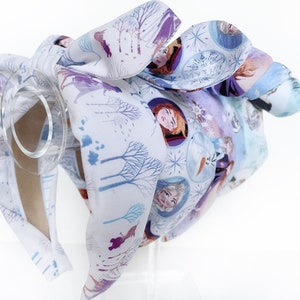May include: A white headband with a purple and blue Frozen print featuring Anna, Elsa, and Olaf. The headband is tied in a bow at the top.