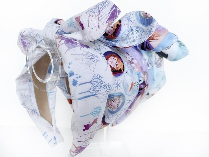 May include: A white headband with a purple and blue Frozen print featuring Anna, Elsa, and Olaf. The headband is tied in a bow at the top.