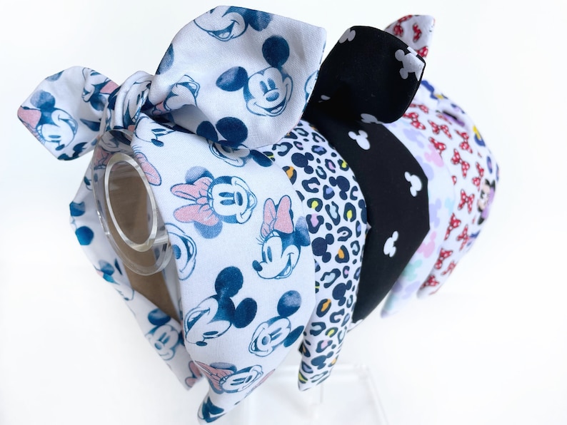 K&ouml;nnte beinhalten: Ein Set aus f&uuml;nf Haarb&auml;ndern mit verschiedenen Mustern. Die Haarb&auml;nder sind aus Stoff und haben oben eine Schleife. Die Muster umfassen einen wei&szlig;en Hintergrund mit blauen und rosa Mickey Mouse-Figuren, einen schwarzen Hintergrund mit wei&szlig;en Mickey Mouse-Figuren, einen Leopardenmuster, einen wei&szlig;en Hintergrund mit rosa und lila Mickey Mouse-Figuren und einen roten Hintergrund mit wei&szlig;en Mickey Mouse-Figuren.