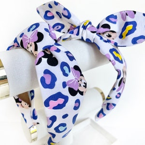 K&ouml;nnte beinhalten: Ein wei&szlig;es Stirnband mit einem blauen, rosa und gelben Leopardenmuster und einem sich wiederholenden Bild von Minnie Mouse. Das Stirnband ist oben zu einer Schleife gebunden.