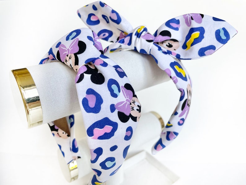 K&ouml;nnte beinhalten: Ein wei&szlig;es Stirnband mit einem blauen, rosa und gelben Leopardenmuster und einem sich wiederholenden Bild von Minnie Mouse. Das Stirnband ist oben zu einer Schleife gebunden.