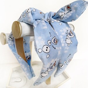 May include: A light blue headband with a repeating pattern of Olaf from Frozen. The headband is tied in a bow at the top.
