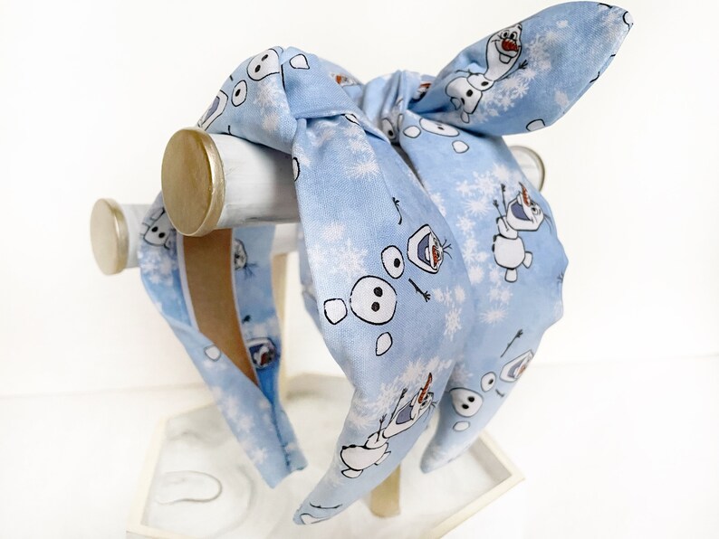 May include: A light blue headband with a repeating pattern of Olaf from Frozen. The headband is tied in a bow at the top.