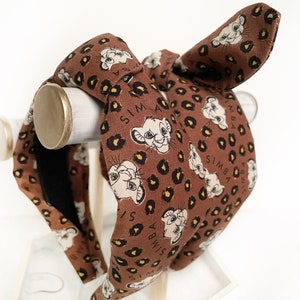 May include: A brown headband featuring a Simba print with a leopard pattern. The headband has a knotted design and a bow detail. The word "SIMBA" is printed on the headband. The headband is displayed on a white stand.
