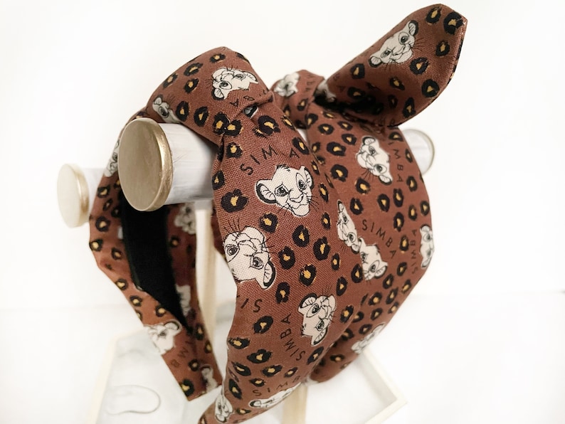 May include: A brown headband featuring a Simba print with a leopard pattern. The headband has a knotted design and a bow detail. The word "SIMBA" is printed on the headband. The headband is displayed on a white stand.