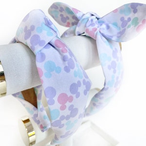 K&ouml;nnte beinhalten: Zwei gebl&uuml;mte Stirnb&auml;nder in Lila, Blau und Rosa mit jeweils einer Schleife. Die Stirnb&auml;nder befinden sich auf einem wei&szlig;en Pr&auml;sentationsst&auml;nder.