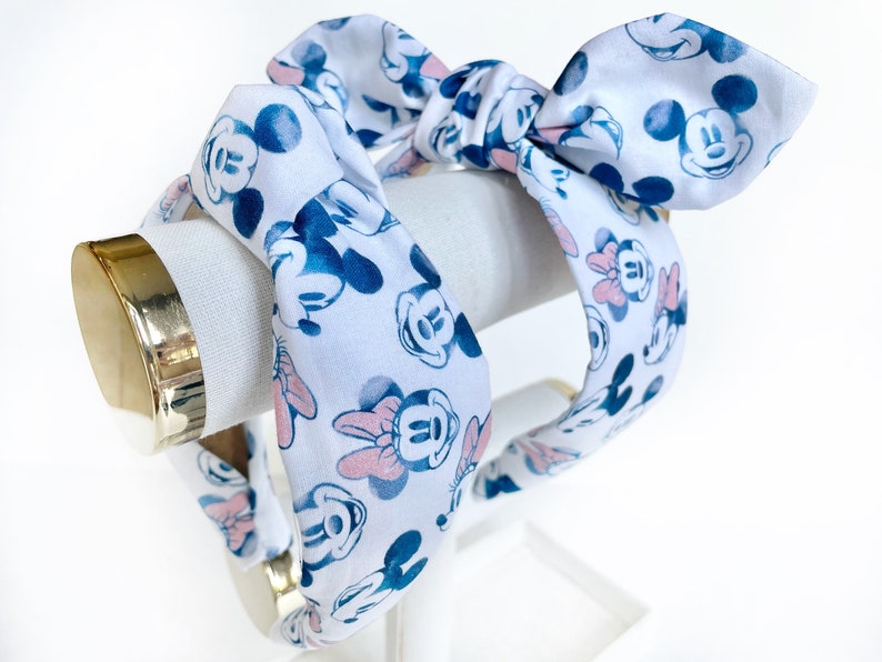 K&ouml;nnte beinhalten: Zwei wei&szlig;e Stirnb&auml;nder mit einem blauen und rosa Minnie Mouse-Druck. Die Stirnb&auml;nder sind zu einer Schleife gebunden.